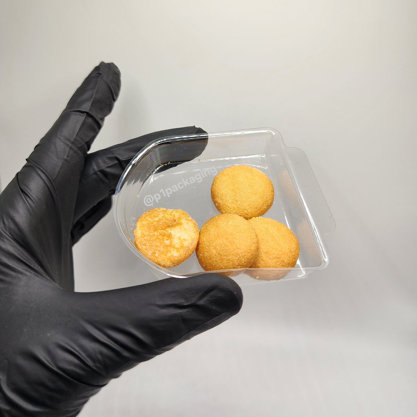 Hand wearing a black glove holding a clear plastic tray with four cookies in it on a white background