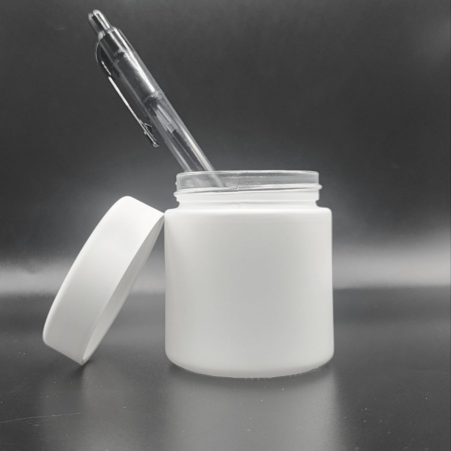 White jar with a white lid and a clear pen inside on a reflective surface with a dark background