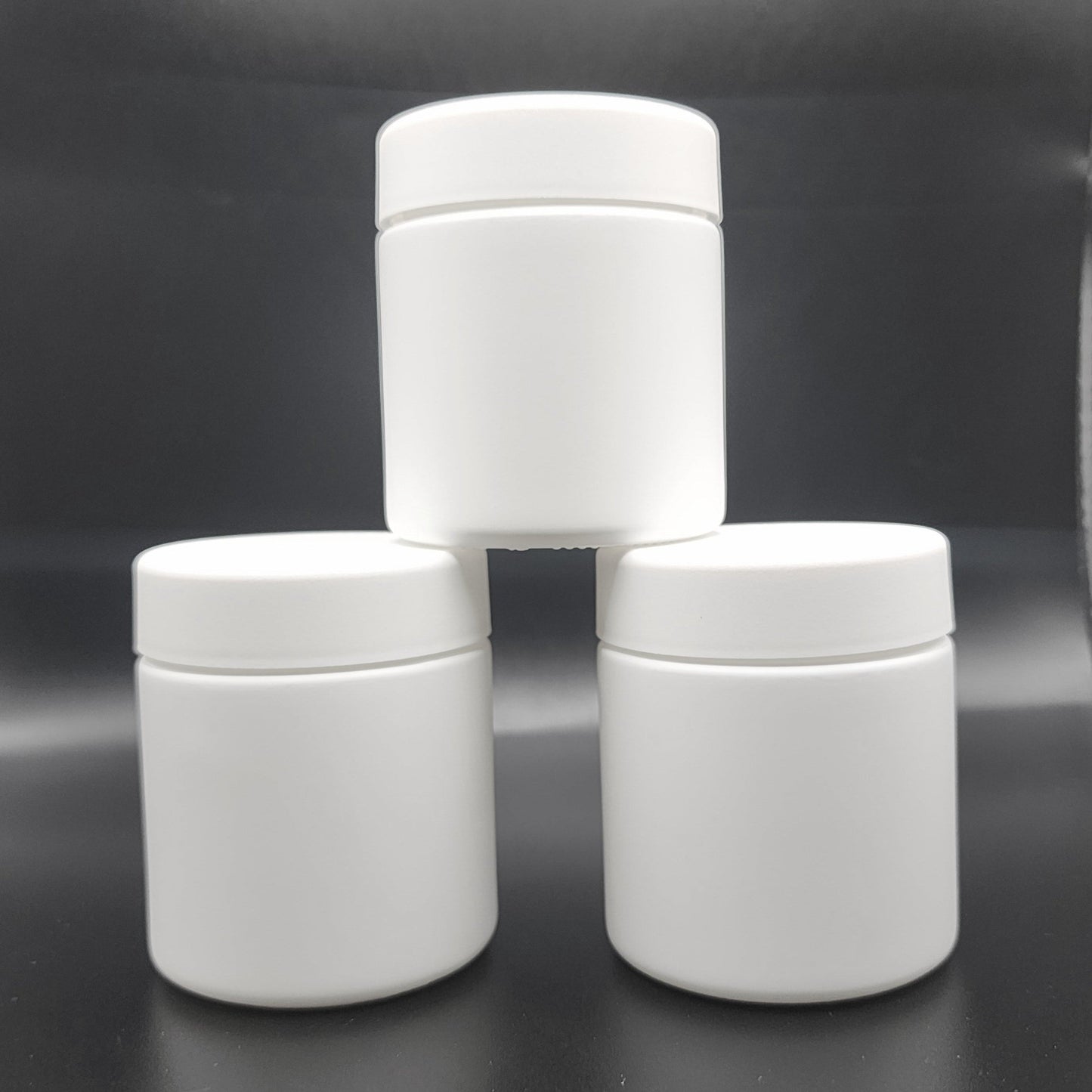 Three white cylindrical jars stacked on a reflective black surface with a blurred background