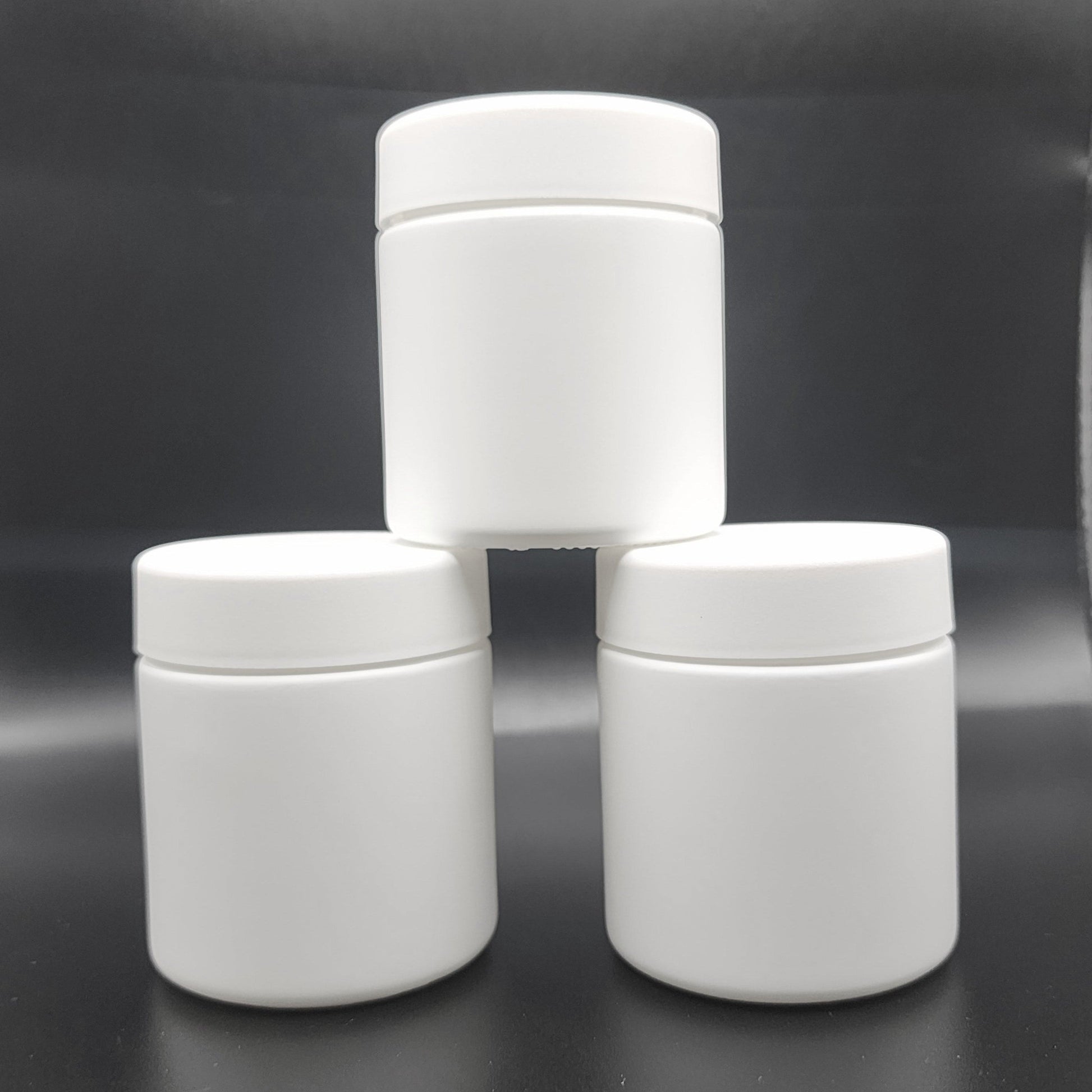 Three white cylindrical jars stacked on a reflective black surface with a blurred background